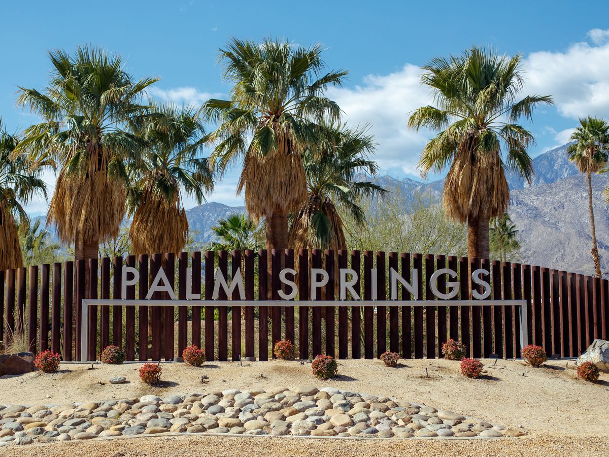 PALM SPRINGS sign in front of palm trees, desert rocks, and mountains under a blue sky