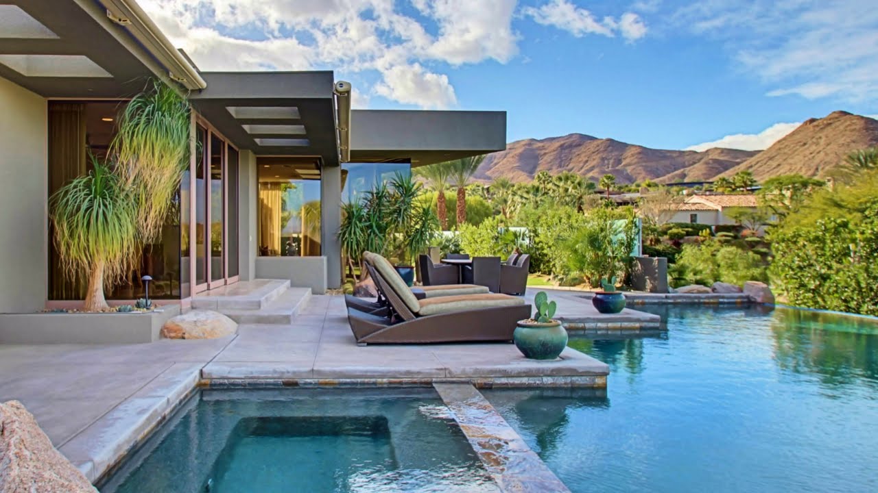 Modern desert villa with infinity pool, spa, and lounge chairs overlooking mountains and lush greenery